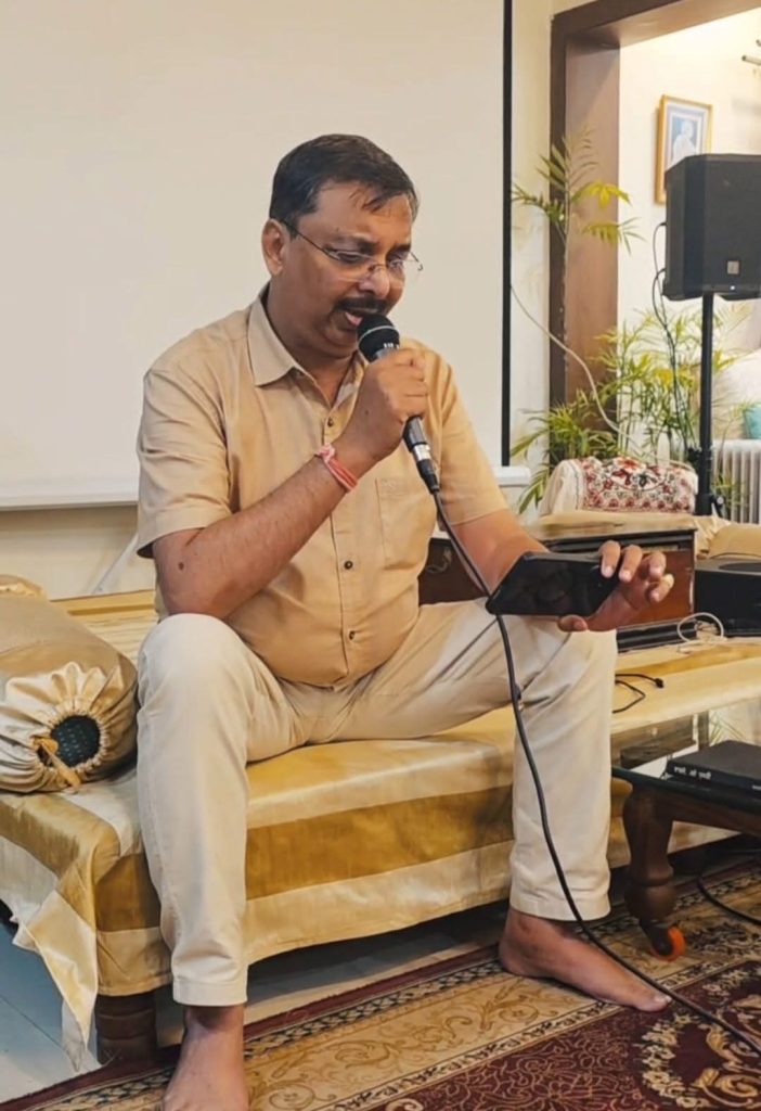 Man in a beige shirt and pants sits cross‑legged on a sofa, speaking into a handheld microphone while looking at his phone indoors at a home gathering.