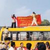 Police officers stand on a yellow bus as protesters raise a red banner on its roof, while a crowd watches nearby.