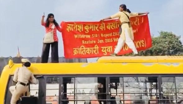 Police officers stand on a yellow bus as protesters raise a red banner on its roof, while a crowd watches nearby.