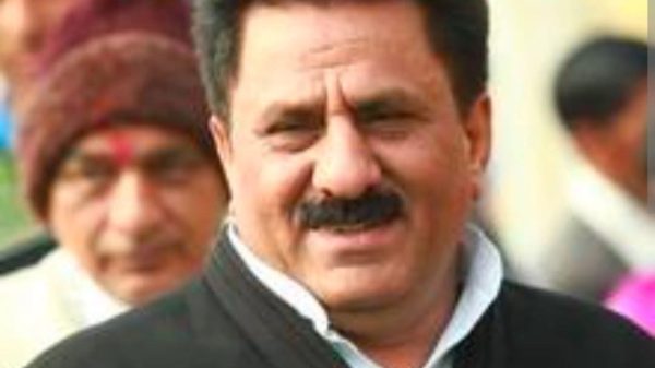 Close-up of a man with dark hair and mustache, wearing a white shirt and black vest, smiling at the camera.
