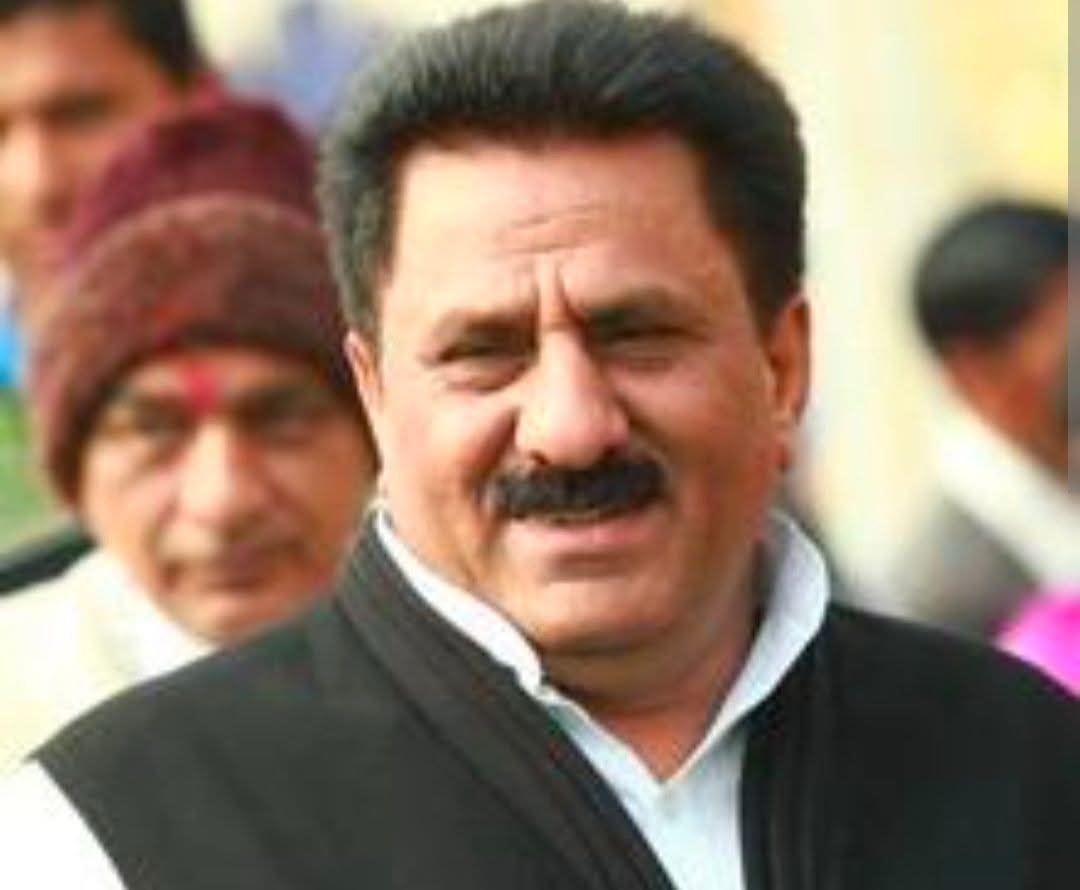 Close-up of a man with dark hair and mustache, wearing a white shirt and black vest, smiling at the camera.