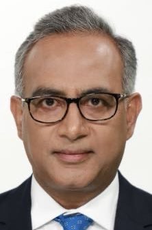 Head-and-shoulders portrait of a middle‑aged man with gray hair and glasses, wearing a dark suit and blue tie, neutral expression.