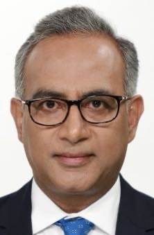 Head-and-shoulders portrait of a middle‑aged man with gray hair and glasses, wearing a dark suit and blue tie, neutral expression.