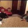 Two men sit across from each other at a floral tablecloth, in a luxurious room, seeming to discuss something.