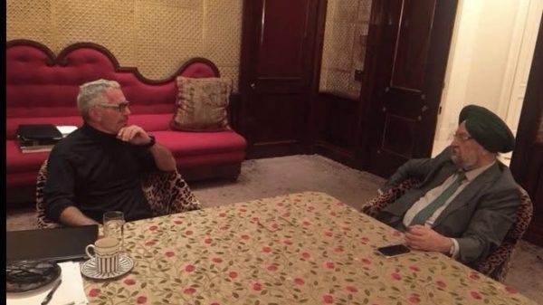 Two men sit across from each other at a floral tablecloth, in a luxurious room, seeming to discuss something.