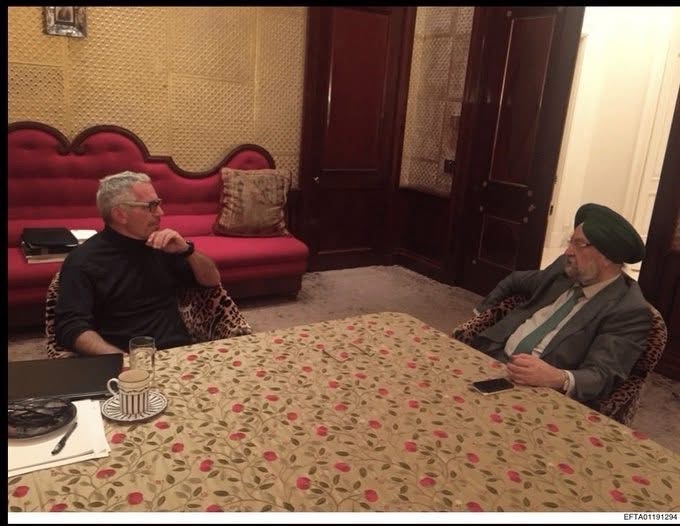 Two men sit across from each other at a floral tablecloth, in a luxurious room, seeming to discuss something.