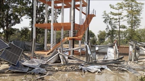 Construction site with bent orange stairwell and scattered metal sheets and debris after a collapse; solar panels lie on the ground.