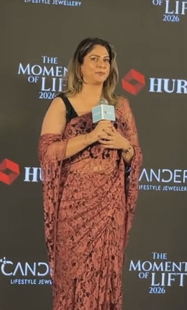 Screenshot | No. 1 Indian Media News Portal Woman in a pink lace saree holding a microphone on a stage with a dark backdrop showing event logos.
