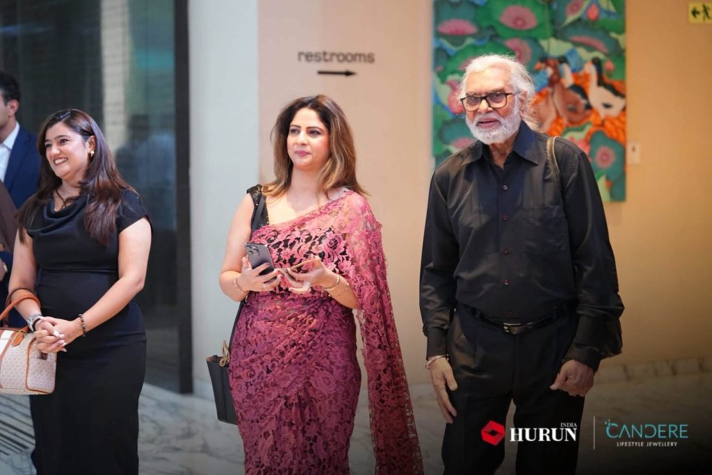 img_2394 | No. 1 Indian Media News Portal Three adults stand indoors at an event: a smiling woman in a black dress on the left, a woman in a pink lace saree holding a phone in the center, and an older man in a black shirt on the right, with a restroom sign and colorful mural in the background.