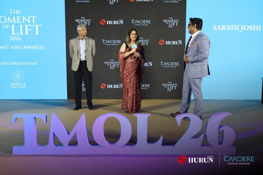 img_2396 | No. 1 Indian Media News Portal Speakers on stage at The Moment of Lift 2026, with a woman in the center holding a microphone and two men beside her.