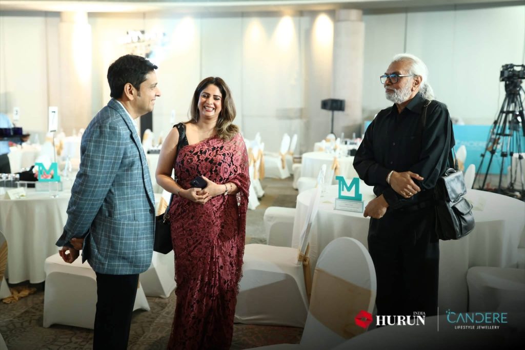 img_2399 | No. 1 Indian Media News Portal Three adults in a banquet hall chat and smile during a formal event, with round tables and a camera setup in the background for a jewelry launch or reception at Hurun/Candere.
