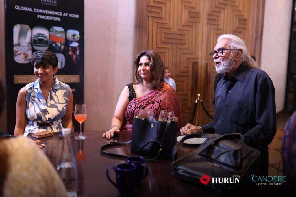 img_2401 | No. 1 Indian Media News Portal Three people standing around a table with drinks, conversing at an indoor event: woman in blue patterned dress left, woman in pink saree center, man with white hair and glasses right; event logos visible bottom right.