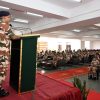 Uniformed officer in camouflage speaks from a green podium to rows of soldiers in a conference hall.