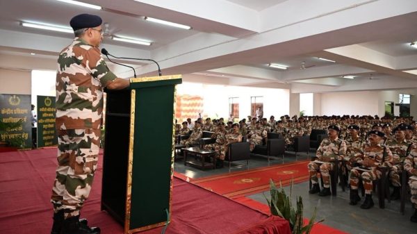 Uniformed officer in camouflage speaks from a green podium to rows of soldiers in a conference hall.