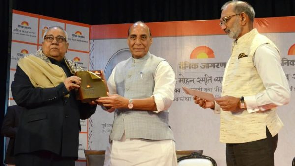 Man in light gray vest presents a bronze plaque to another man in a dark suit at an awards ceremony, with a third man reading materials nearby.