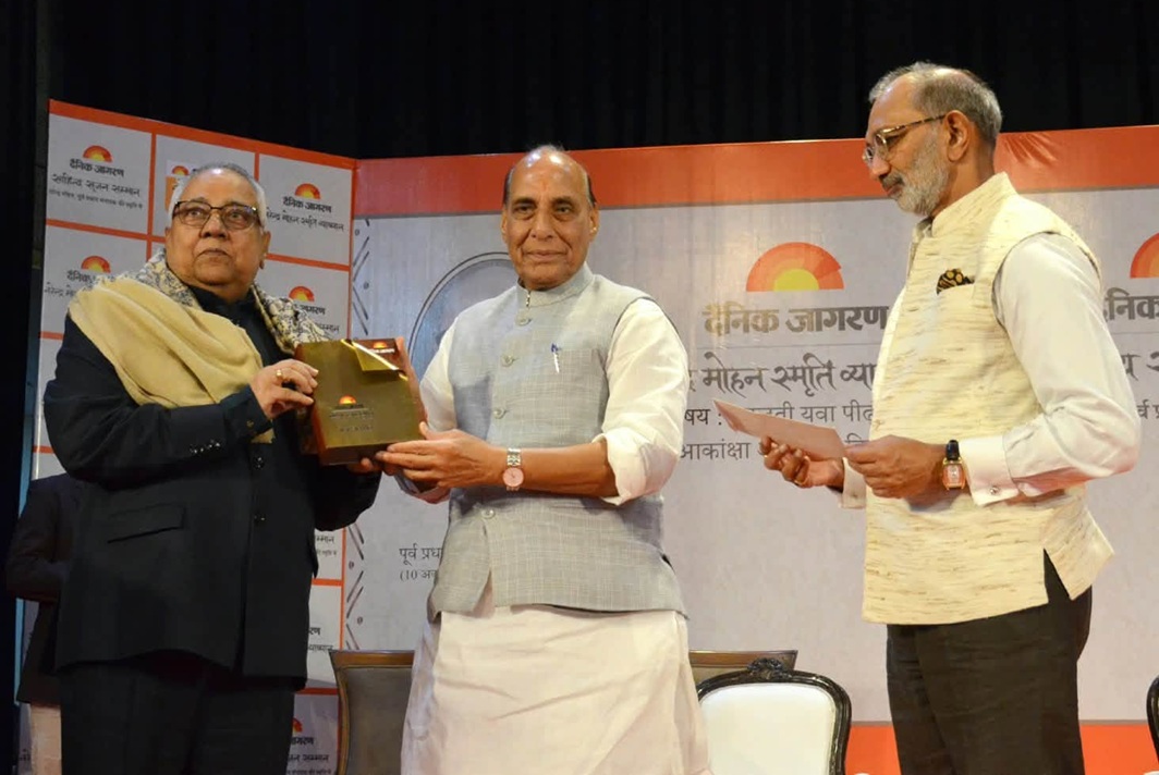 jagran sahitya | No. 1 Indian Media News Portal Man in light gray vest presents a bronze plaque to another man in a dark suit at an awards ceremony, with a third man reading materials nearby.