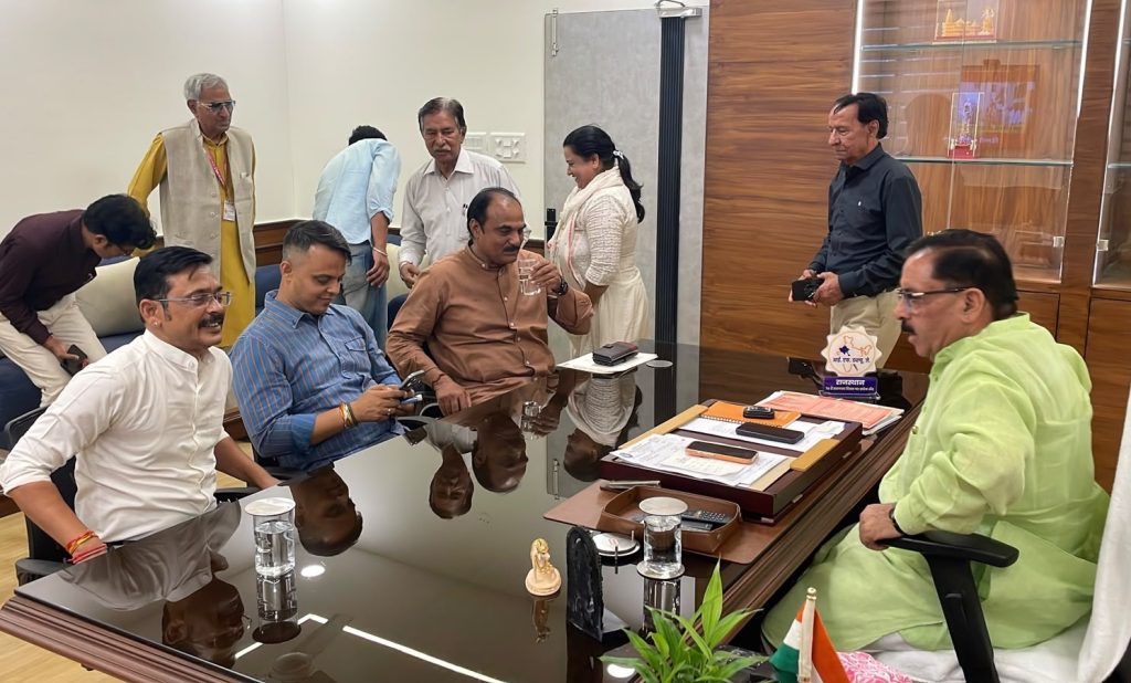 jaipur_1 | No. 1 Indian Media News Portal Senior man in a bright lime-green kurta sits at a large glass desk, surrounded by colleagues chatting and checking phones in a sunny office.