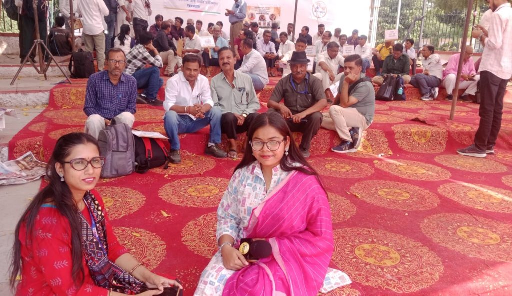 jaipur_2 | No. 1 Indian Media News Portal Two women in bright sarees sit on a red patterned carpet at a crowded outdoor gathering, with many people seated in the background.