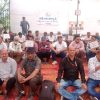 Group of men seated on a red carpet at an outdoor gathering, some holding placards while a speaker addresses from a small stage in the background.