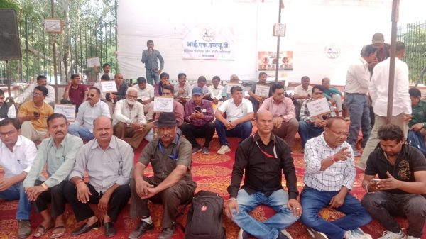 Group of men seated on a red carpet at an outdoor gathering, some holding placards while a speaker addresses from a small stage in the background.