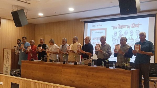 Group of men standing along a table on a stage, each holding a book during a book-launch event, with a projector screen in the background.