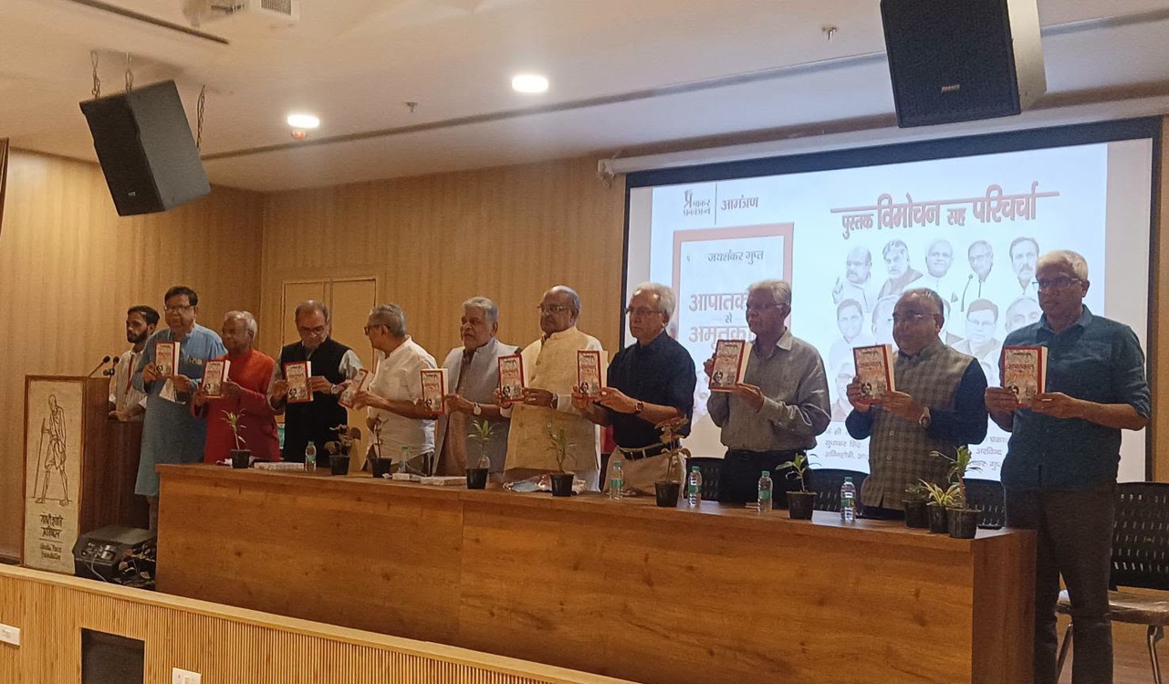 Group of men standing along a table on a stage, each holding a book during a book-launch event, with a projector screen in the background.