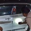 Two people inspect a white SUV at night; one in uniform shines a light at the driver's door.