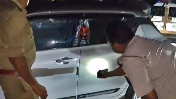 Two people inspect a white SUV at night; one in uniform shines a light at the driver's door.