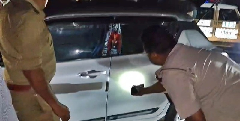 Two people inspect a white SUV at night; one in uniform shines a light at the driver's door.