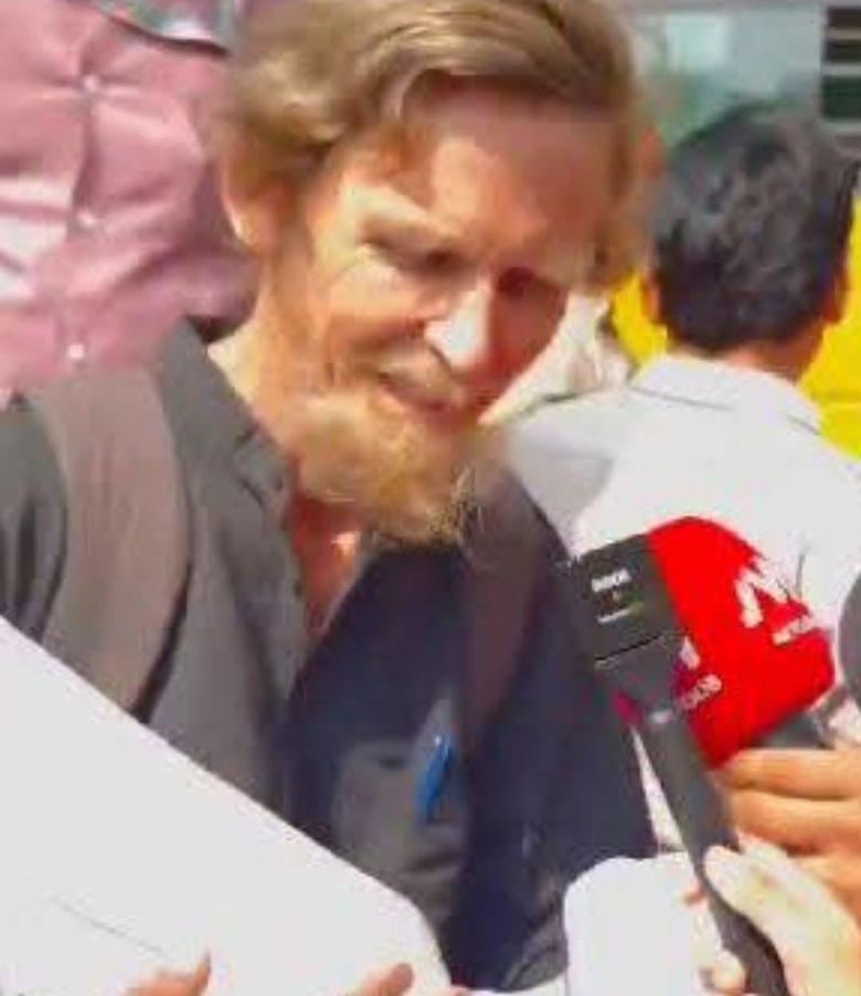 Bearded man in a dark blazer being interviewed by reporters holding a red microphone outdoors.
