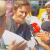 Bearded man in a dark shirt speaks to reporters, several microphones held toward him as a crowd and police officers look on.