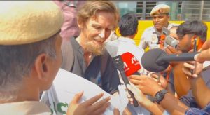 Bearded man in a dark shirt speaks to reporters, several microphones held toward him as a crowd and police officers look on.