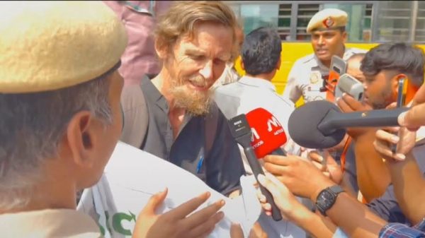 Bearded man in a dark shirt speaks to reporters, several microphones held toward him as a crowd and police officers look on.