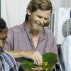 Smiling man with a beard and plaid shirt eats a meal served on a large green banana leaf plate.