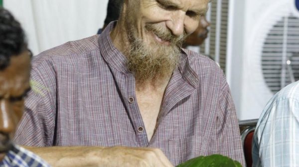 Smiling man with a beard and plaid shirt eats a meal served on a large green banana leaf plate.