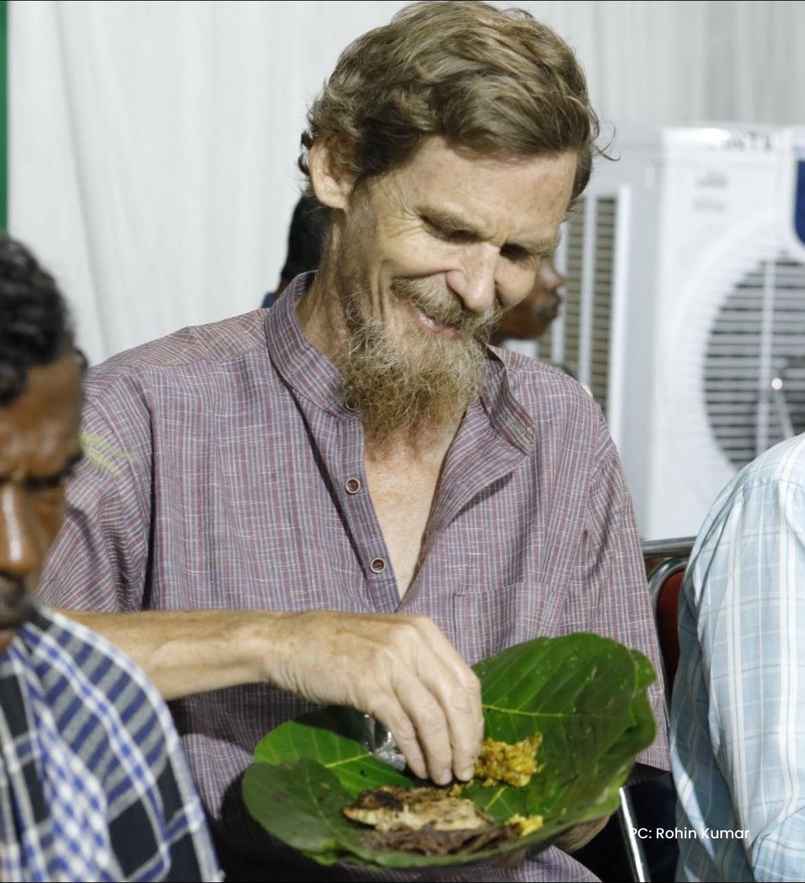 Smiling man with a beard and plaid shirt eats a meal served on a large green banana leaf plate.