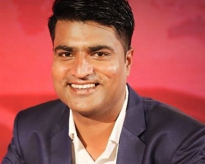 Man in a navy suit and white shirt smiling at the camera with a red backdrop behind him.