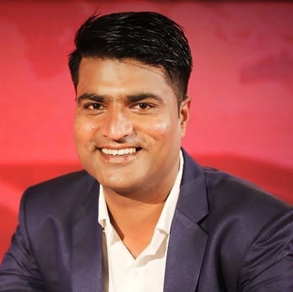 jitendra shukla | No. 1 Indian Media News Portal Man in a navy suit and white shirt smiling at the camera with a red backdrop behind him.
