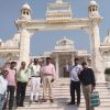 Seven men pose on marble steps in front of a white ornate temple entrance under a clear blue sky, each making a peace sign.