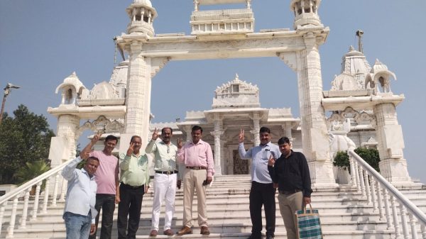 Seven men pose on marble steps in front of a white ornate temple entrance under a clear blue sky, each making a peace sign.