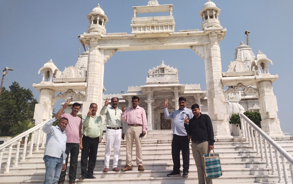 Seven men pose on marble steps in front of a white ornate temple entrance under a clear blue sky, each making a peace sign.