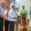 Men stand in a row around a decorated bust on a stage, garlands and lit lamps in front of the statue as a ceremony unfolds enhanced by flower arrangements in the background.