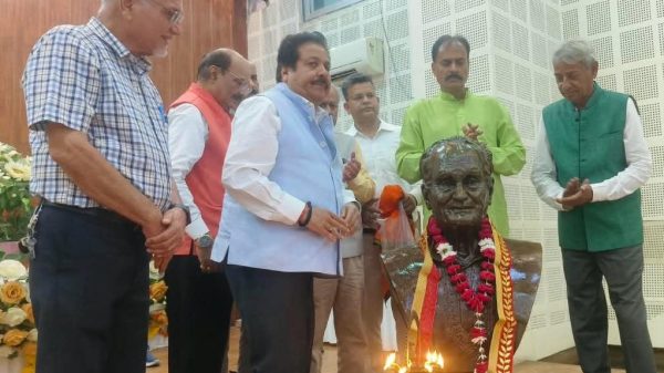 Men stand in a row around a decorated bust on a stage, garlands and lit lamps in front of the statue as a ceremony unfolds enhanced by flower arrangements in the background.