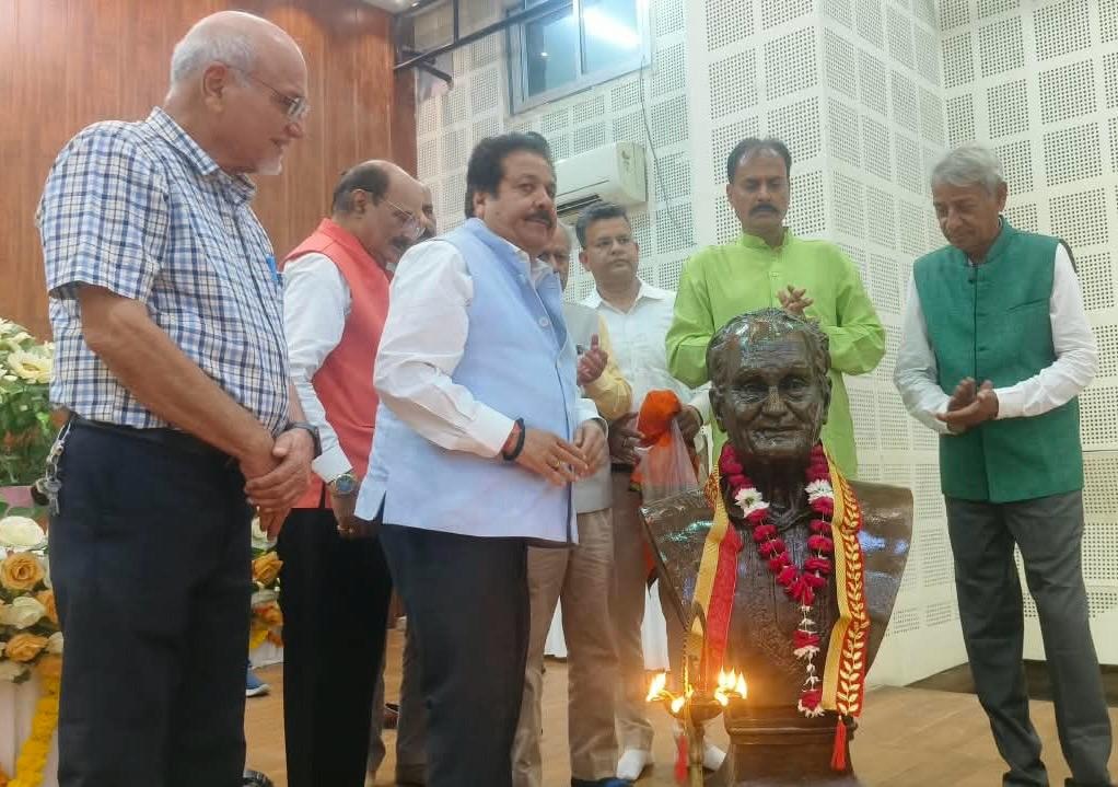Men stand in a row around a decorated bust on a stage, garlands and lit lamps in front of the statue as a ceremony unfolds enhanced by flower arrangements in the background.