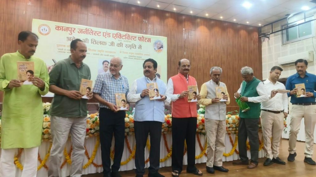 Ten men stand in a row on a stage holding copies of a booklet, at a formal event with a Hindi banner behind them.