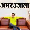 Man in a lime shirt lounging with arms on a brown leather sofa beneath an Amar Ujala sign; a newspaper rack sits to the right.