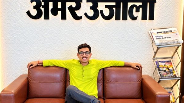Man in a lime shirt lounging with arms on a brown leather sofa beneath an Amar Ujala sign; a newspaper rack sits to the right.