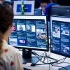Person seated at a desk viewing two large monitors filled with social feeds and news articles; a Brazilian flag is visible in the background.