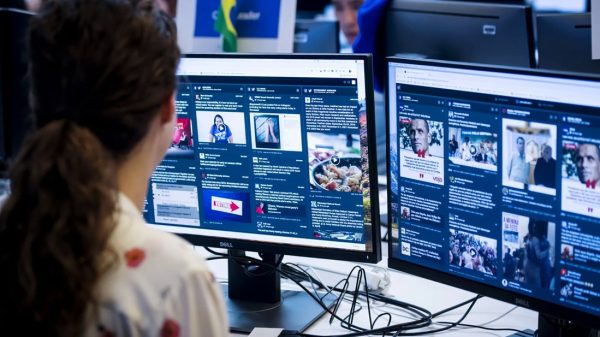 Person seated at a desk viewing two large monitors filled with social feeds and news articles; a Brazilian flag is visible in the background.