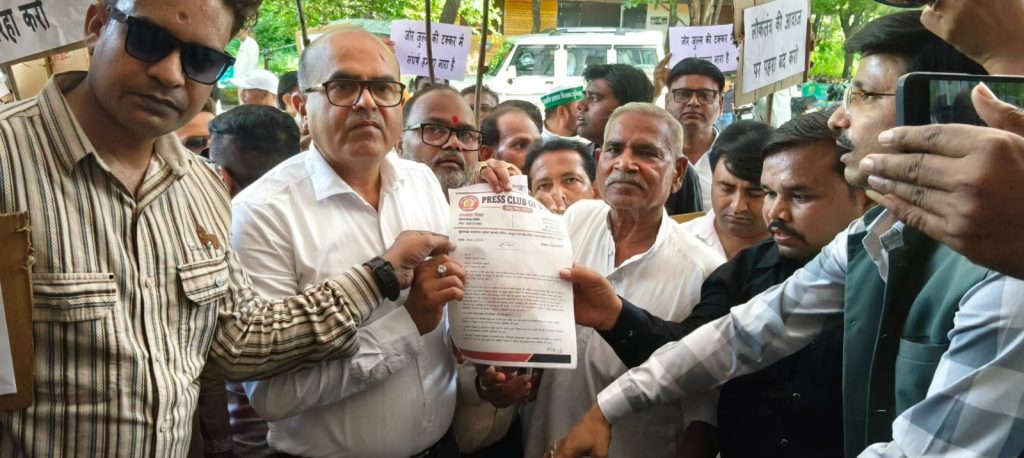 Group of men at a protest handing a printed document to an official amid signs in Hindi in the background.